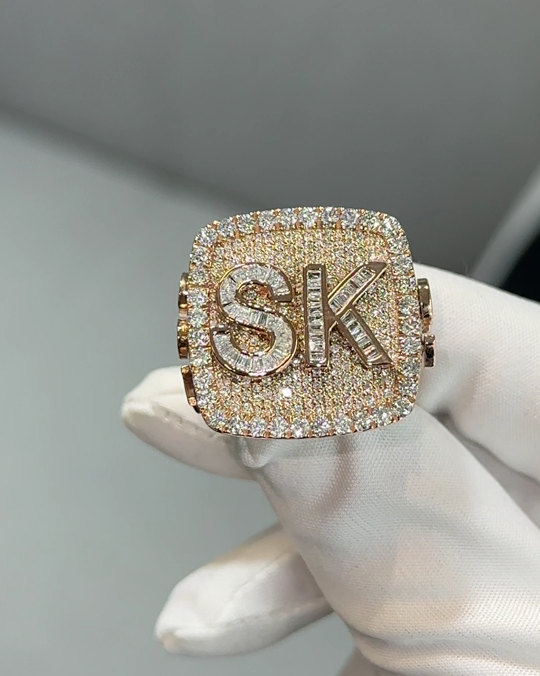 Front view of a rose gold championship-style ring with a diamond-set top and raised diamond-set initials, held in a white-gloved hand